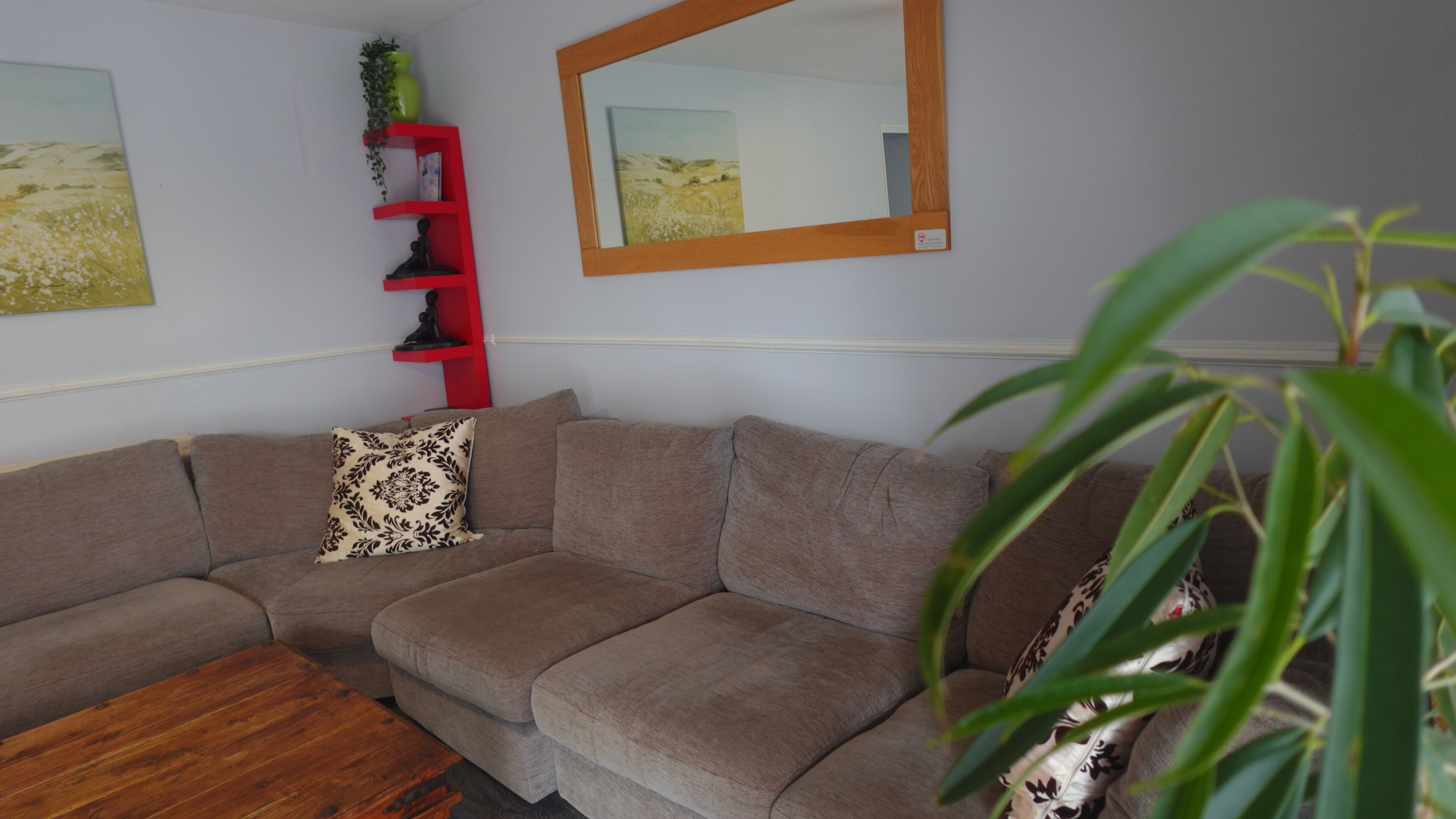 Ein Wohnzimmer mit einem braunen Ecksofa, einem Holztisch, einer roten Regaleinheit mit Dekoration und einem großen Spiegel an der Wand.