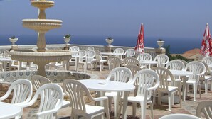 Une terrasse avec des chaises et des tables en plastique blanc, une fontaine en pierre et des parasols rouges.