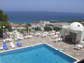 Ein Poolbereich mit Liegestühlen und Sonnenschirmen, eine weiße Kuppelstruktur und Blick auf das Meer.