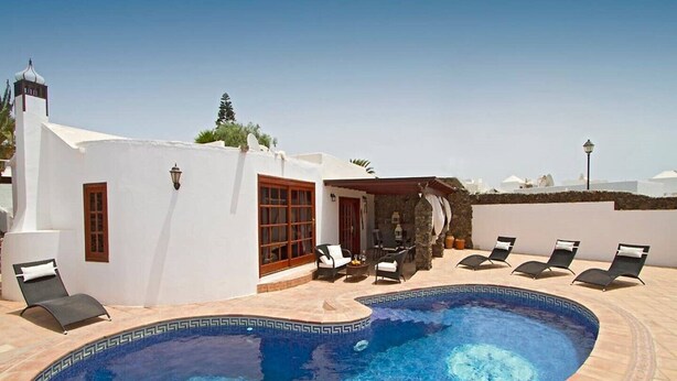 A pool area with lounge chairs, a stone wall, and a white building with a door and windows.