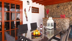 An outdoor dining area with wicker chairs and a glass table, a stone wall, and a wooden ceiling.