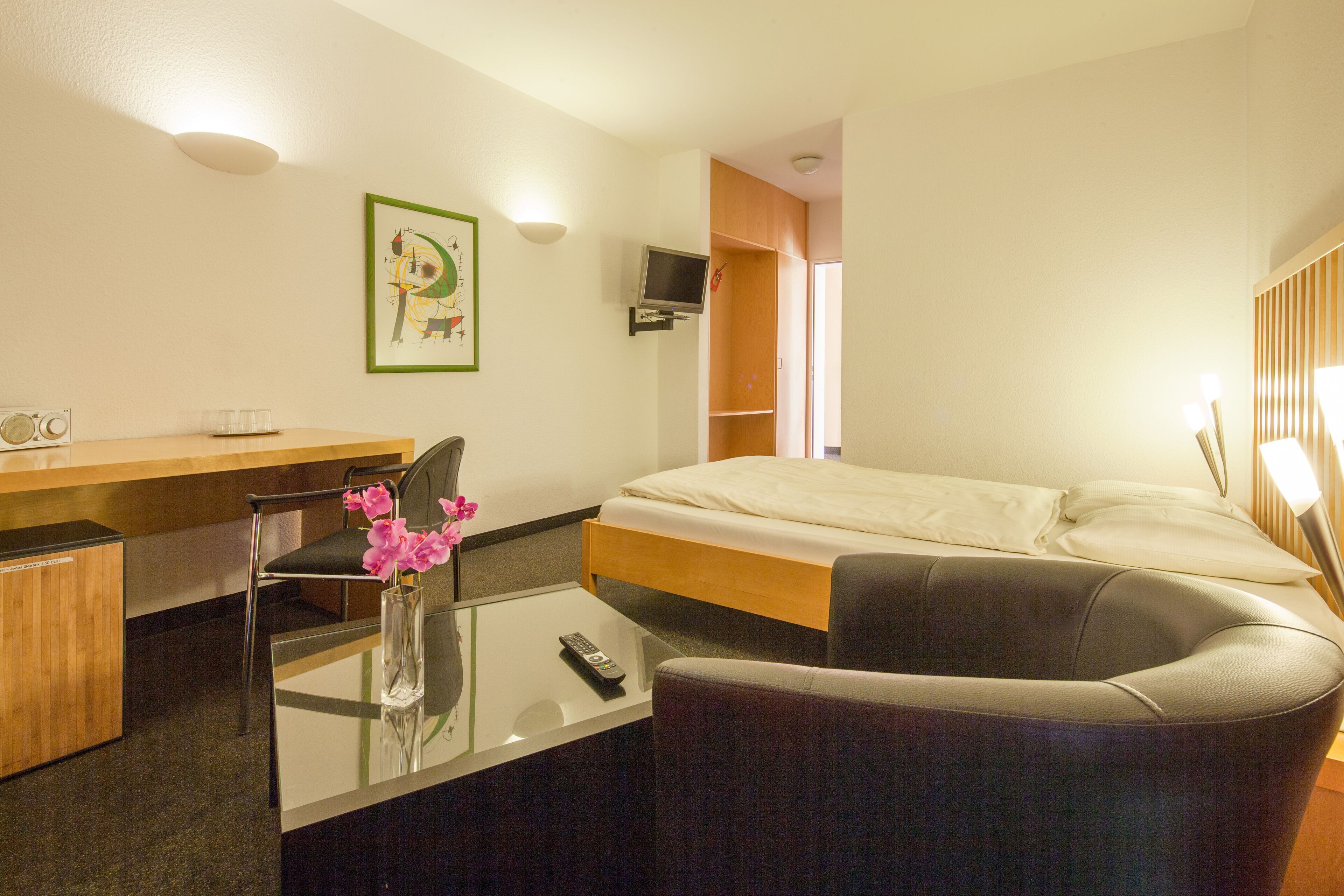 Una habitación de hotel con una cama, un escritorio con una silla, un televisor montado en la pared y una mesita con un jarrón de flores.