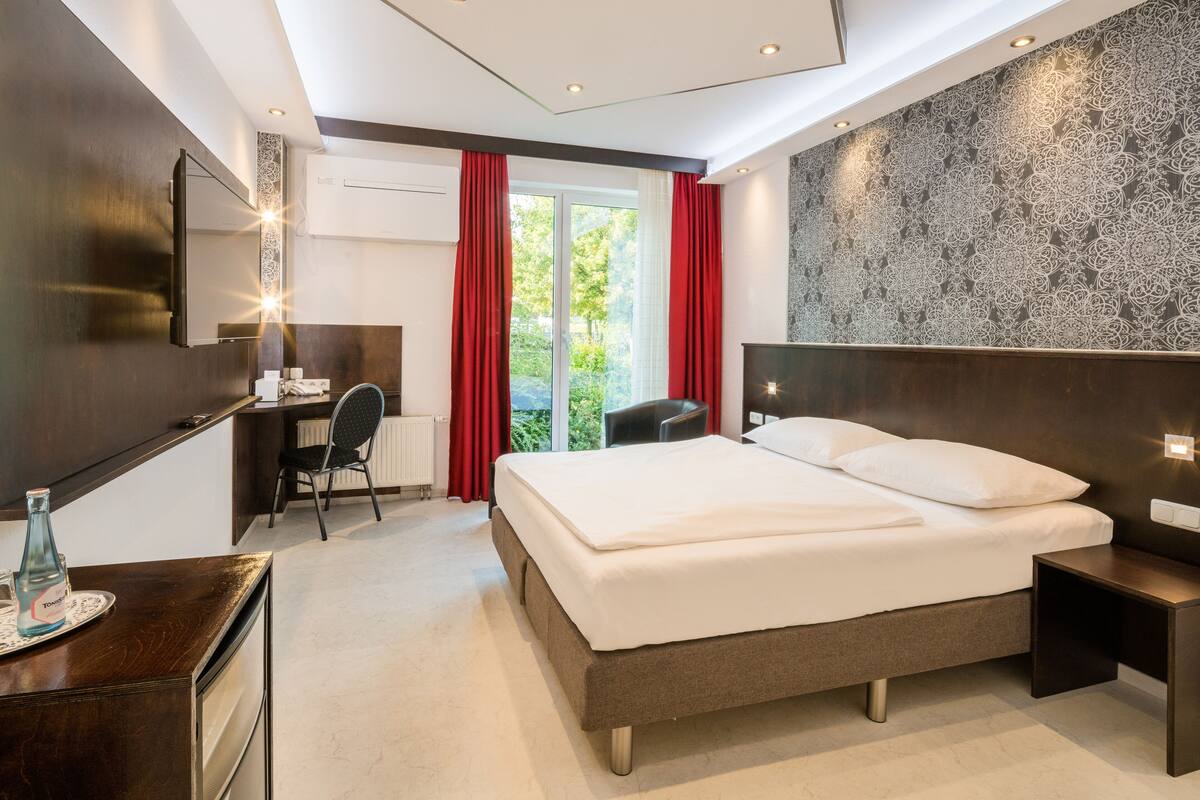 A modern hotel room with a large bed, a desk with a chair, a mirror, and a view of greenery outside.