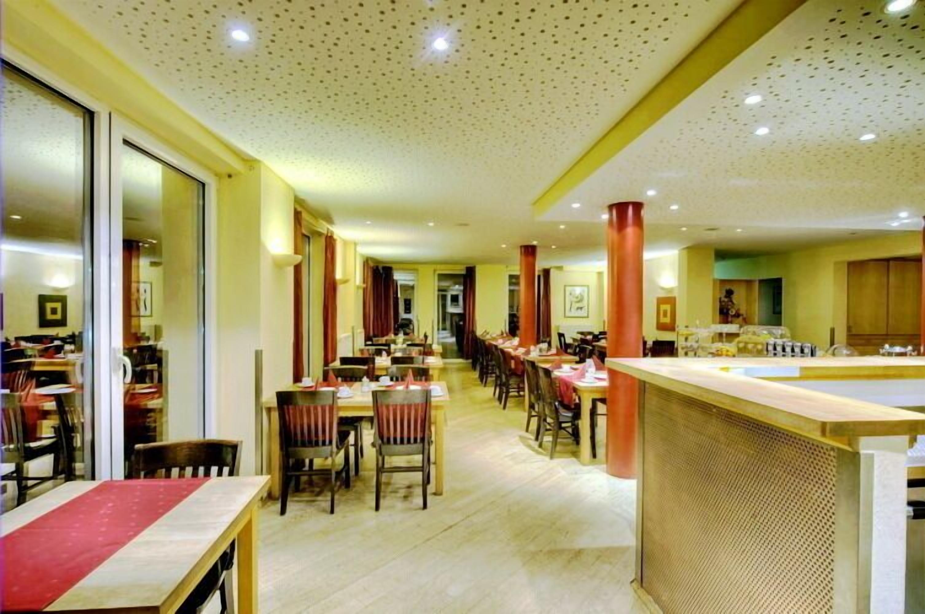 A spacious hotel dining area with multiple tables and chairs, a central bar area, and a long corridor with doors leading to other rooms.