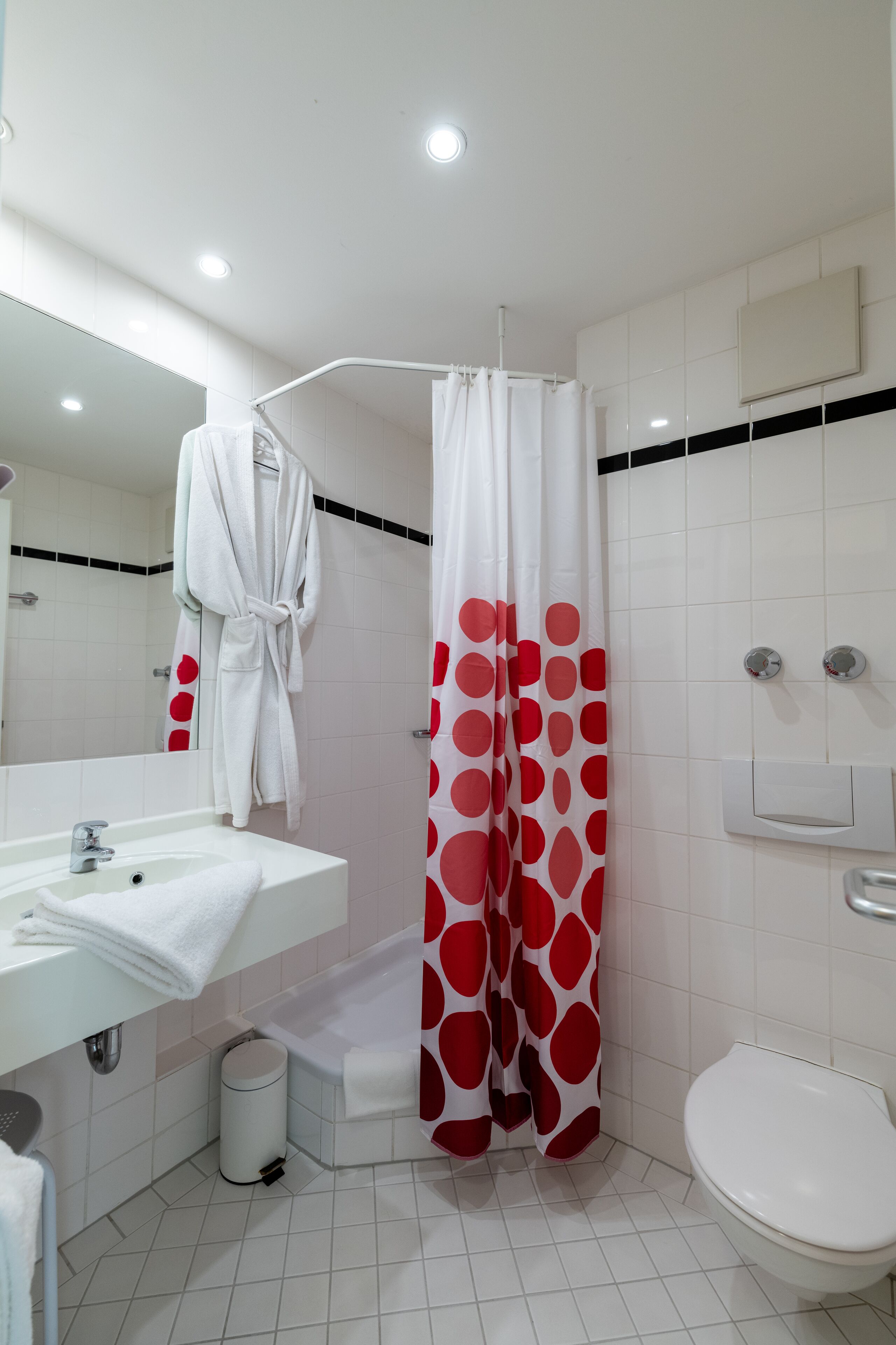 Un baño con un lavabo blanco, un inodoro, una ducha con cortina de lunares rojos y blancos y una bata blanca colgada en la pared.