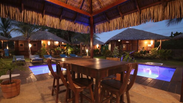 A wooden dining table with chairs, a pool, and thatched-roof structures in a tropical setting. - Atins Charme Chales (Barreirinhas)