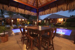 Una mesa de madera con sillas, una piscina y construcciones con techo de paja en un entorno tropical.