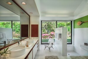 Un baño moderno con un espejo grande, dos lavamanos, una zona de ducha y vistas a un jardín a través de amplios ventanales.
