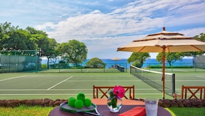 Una cancha de tenis con red, sillas y una sombrilla. Hay una mesa con una raqueta, pelotas, un jarrón con flores y un vaso de agua.