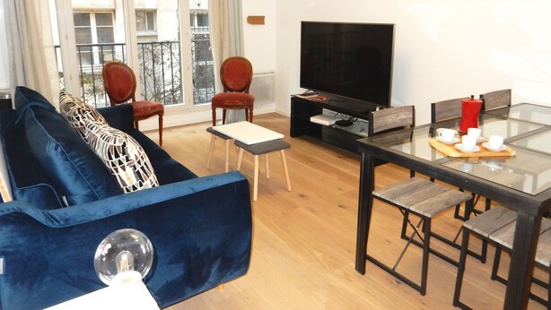 A modern living room with a blue sofa, two red chairs, a glass dining table with four chairs, a flat-screen TV, and a coffee maker.
