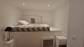 A small, white room with a bunk bed, a bedside lamp, a radiator, and a framed picture on the wall.