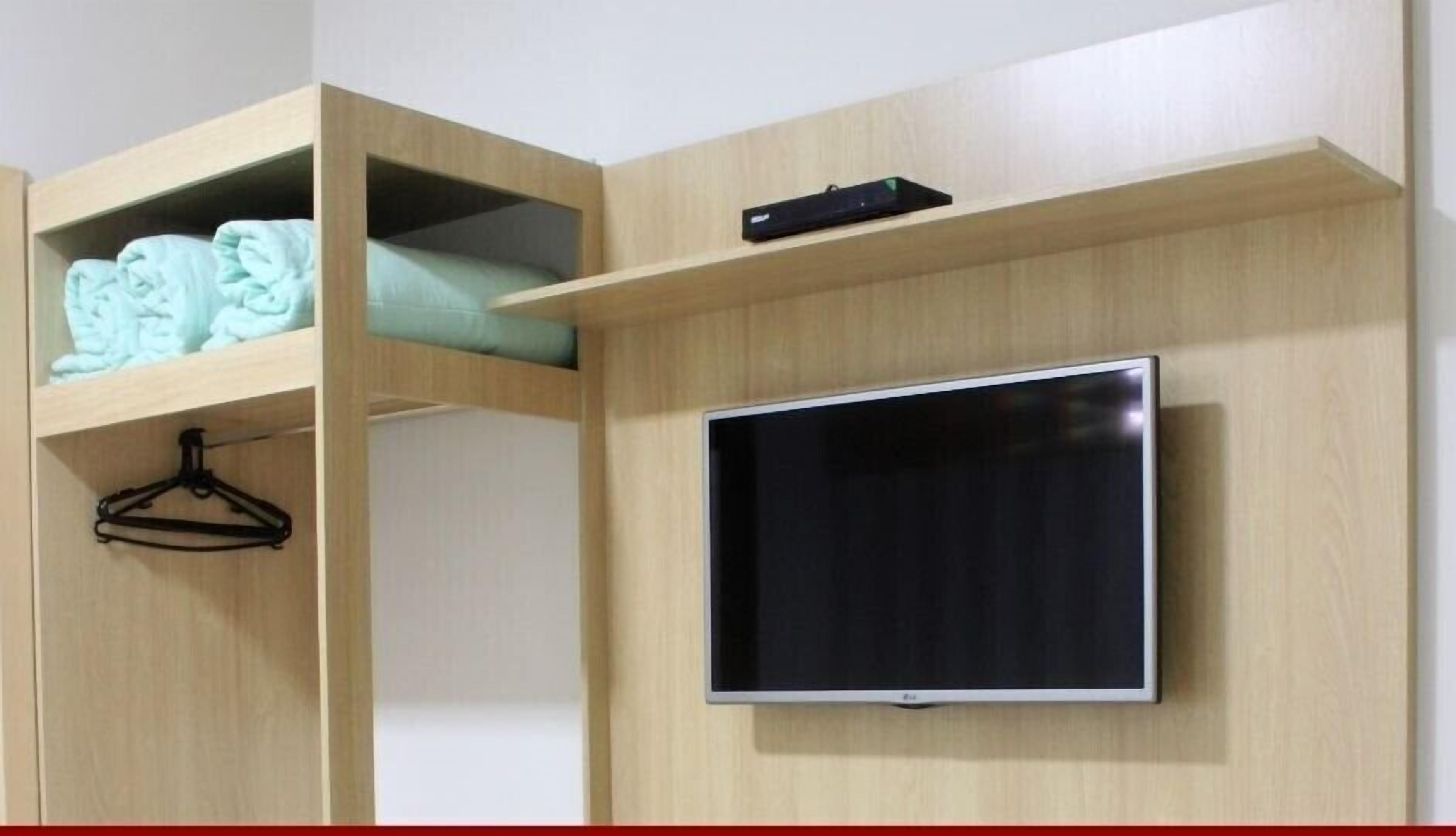 A room with a TV mounted on the wall, a wooden shelf with folded clothes, and a black hanger.