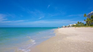 A beach with clear blue water, sandy shore, and a building with palm trees.