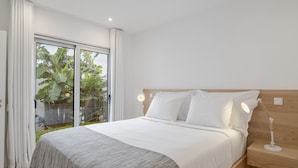 A modern bedroom with a large bed, white bedding, a wooden headboard, a bedside table with a lamp, and a sliding glass door leading to a balcony with plants.