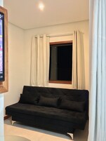 Una habitación con un sofá negro, una ventana con cortinas blancas y un cuadro con marco de madera en la pared.