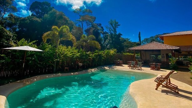 Un hombre descansando en una piscina curvada con agua azul cristalina, rodeado de exuberante vegetación y un cielo soleado.