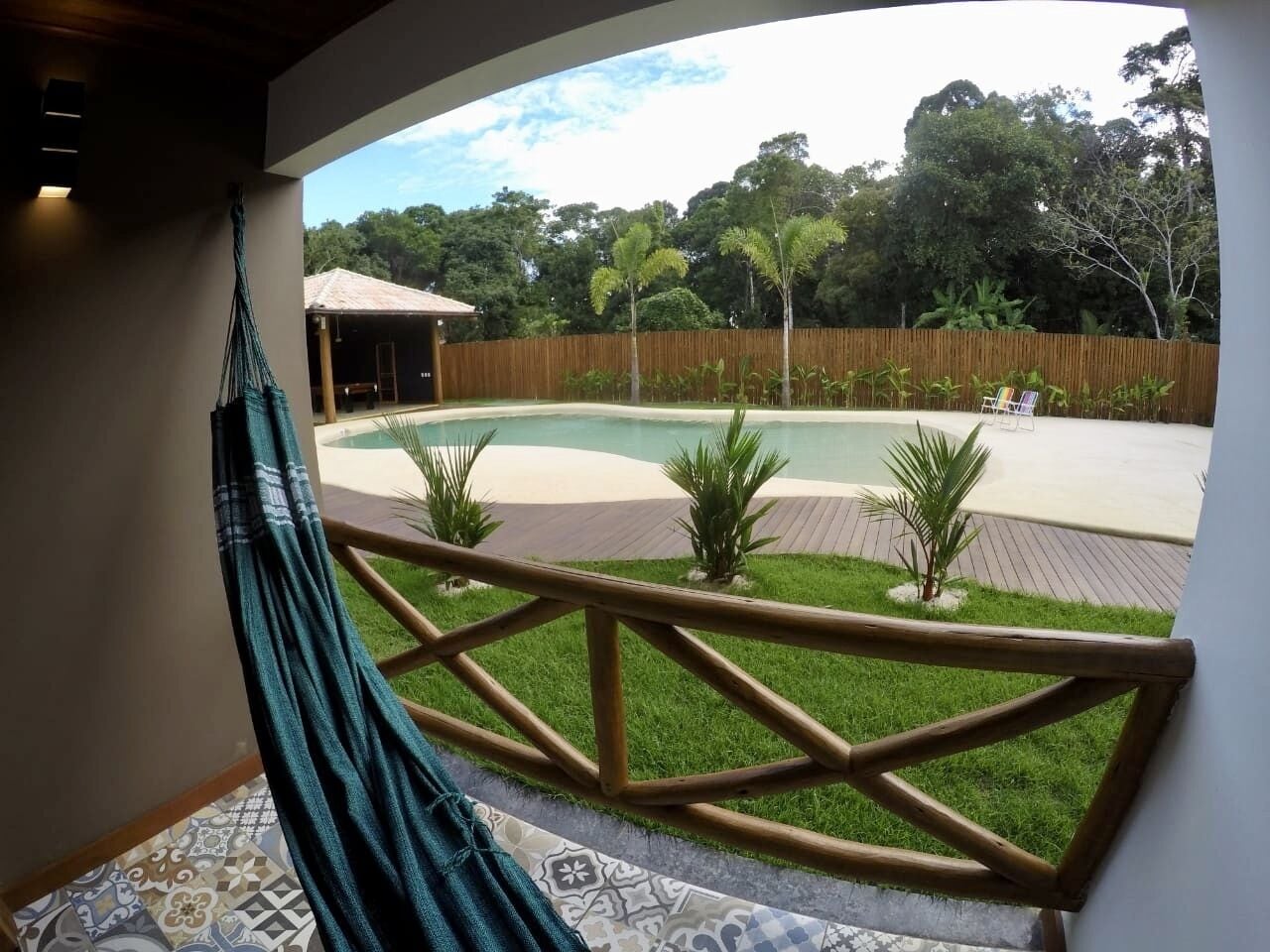 Uma rede numa varanda com vista para uma piscina, cerca de madeira e plantas tropicais.
