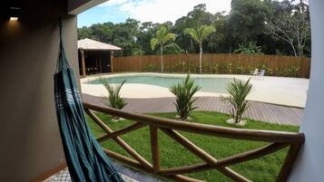 Uma rede numa varanda com vista para uma piscina, cerca de madeira e plantas tropicais.