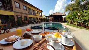 Mesa de desayuno con platos de comida, tazas y una botella, con vista a una piscina, un edificio y palmeras.