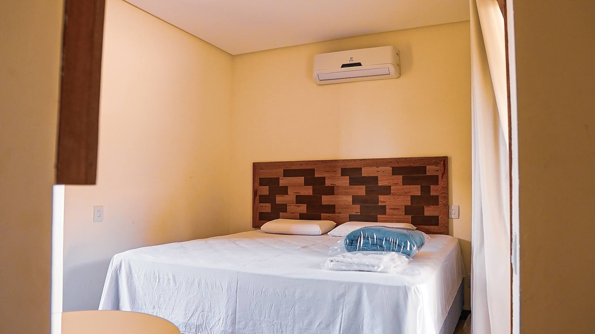 A hotel room with a bed, a wooden headboard, a white bedspread, and a wall-mounted air conditioner.