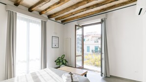 Een slaapkamer met een groot bed, een houten plafond, een raam met gordijnen, een plant en uitzicht op gebouwen.