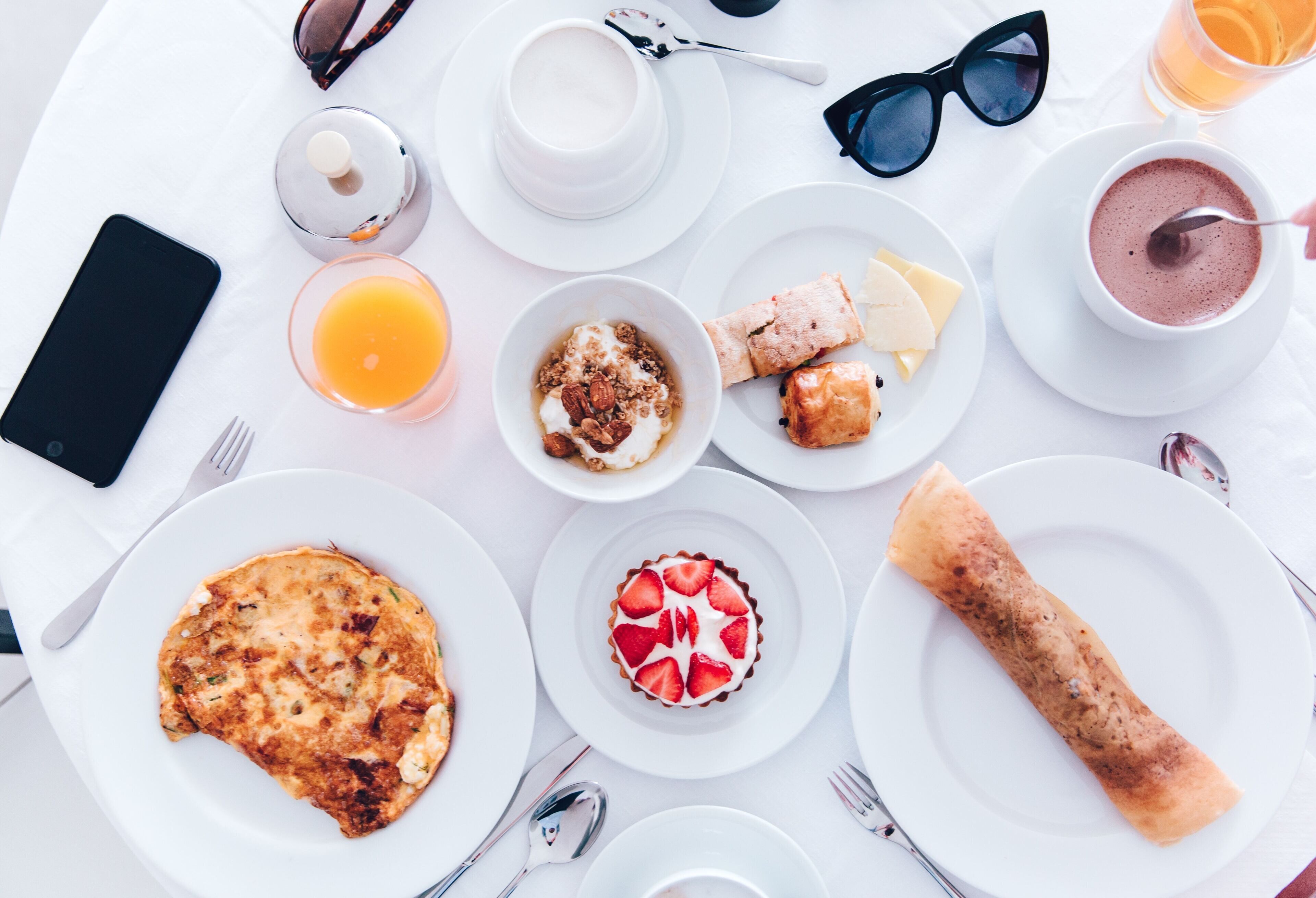Une table blanche dressée pour le petit-déjeuner, avec différents plats, un téléphone intelligent et une tasse de café.