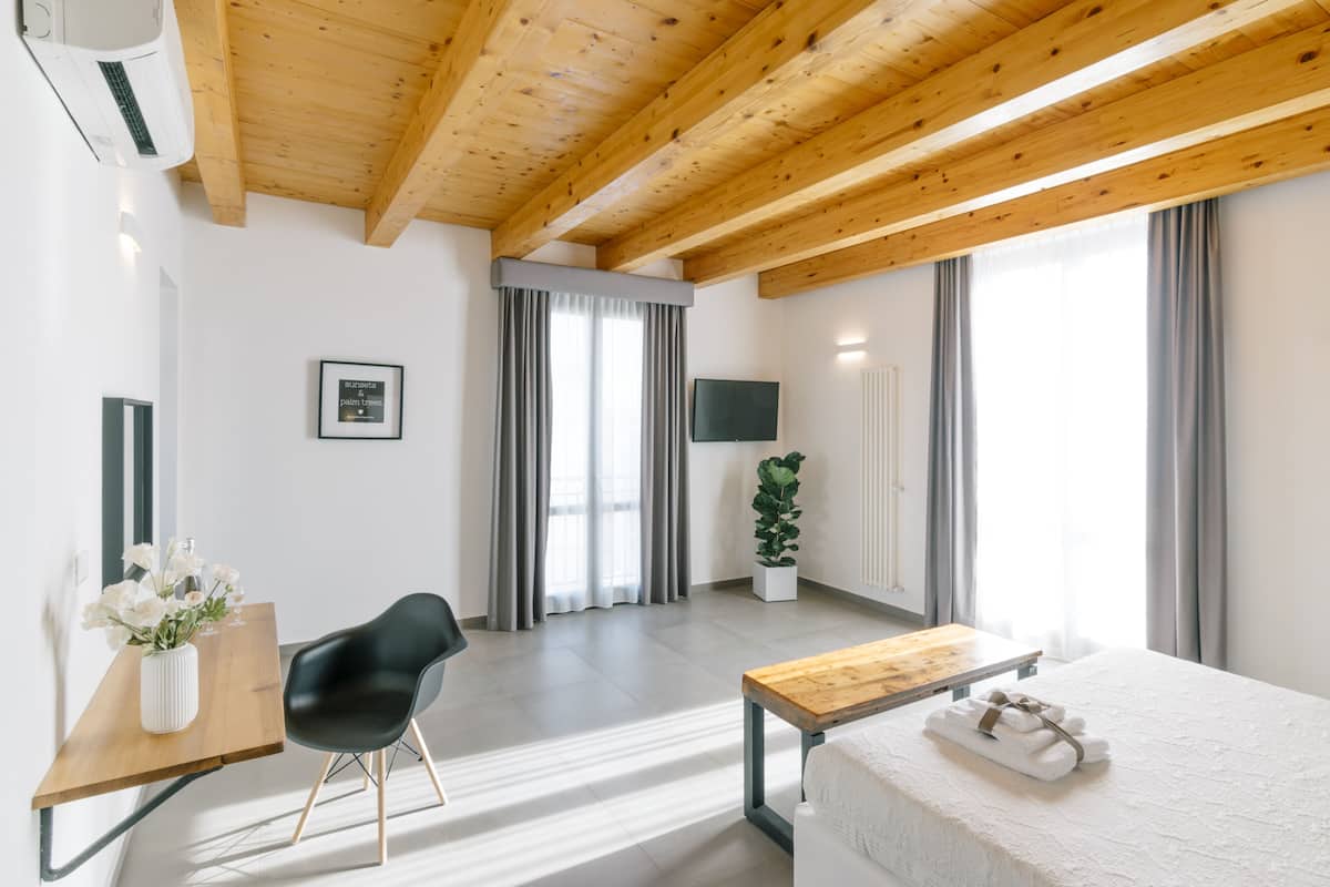 Une chambre d’hôtel moderne avec un plafond en bois, un lit recouvert de draps blancs, un bureau en bois orné d’un vase de fleurs, une chaise noire, une plante en pot et un téléviseur fixé au mur.