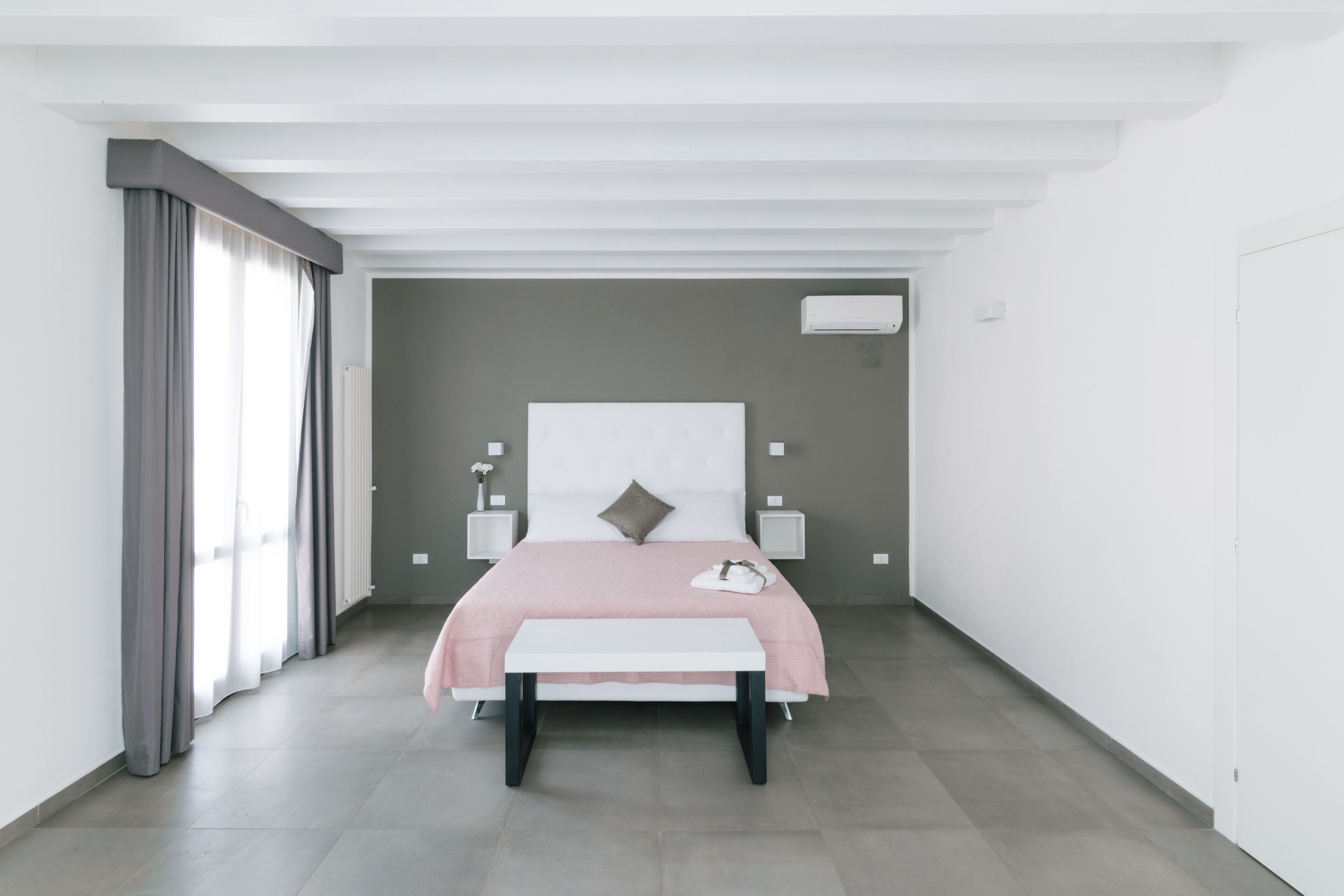 A modern bedroom with a large bed, bedside tables, a bench, and a window with curtains.