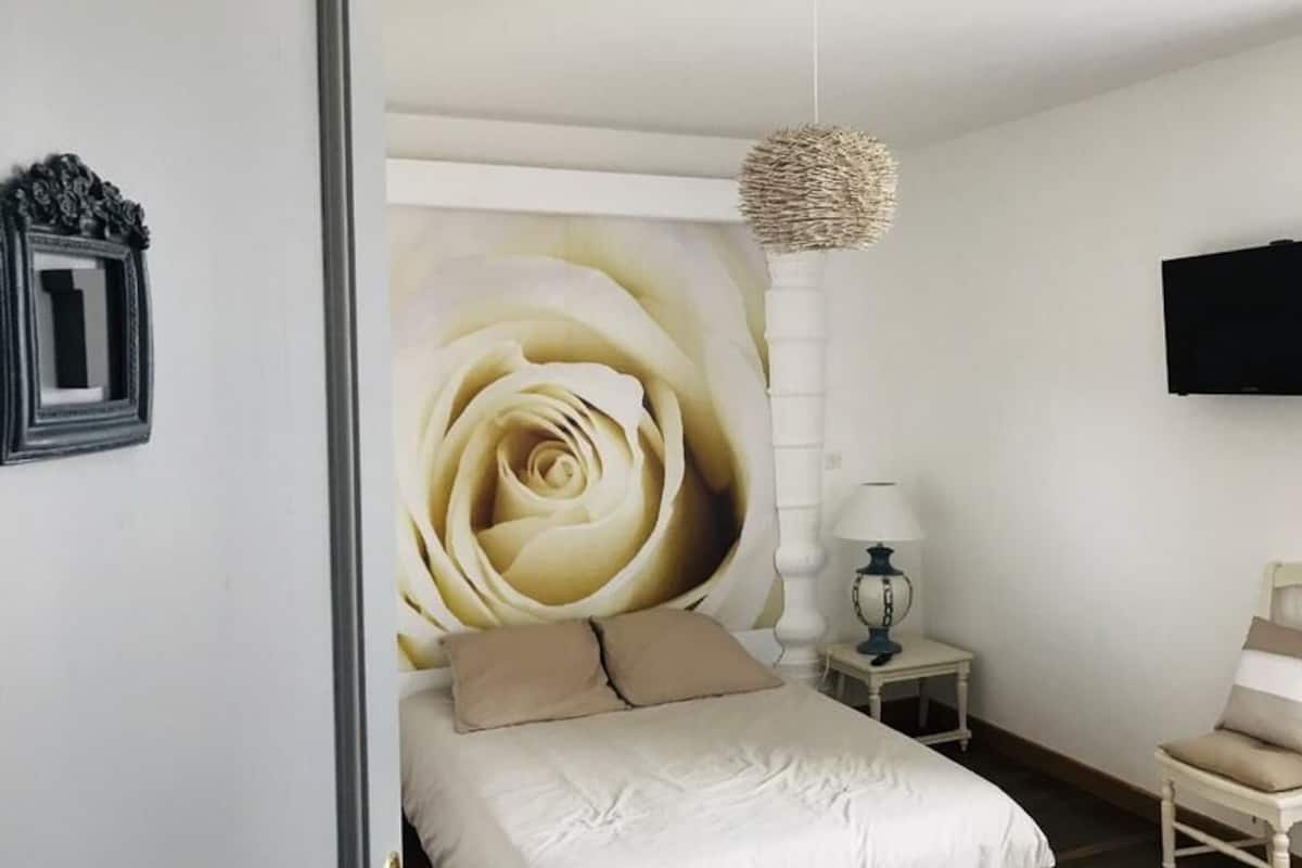 A bedroom with a large wall mural of a rose, a bed with beige bedding, a bedside table with a lamp, a chair, and a flat-screen TV mounted on the wall.