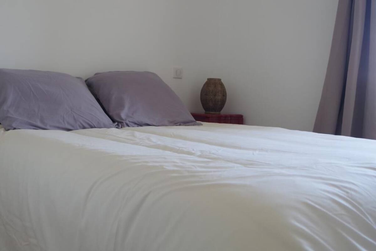 A bed with white and purple bedding, a small wooden nightstand with a vase, and a plain wall.