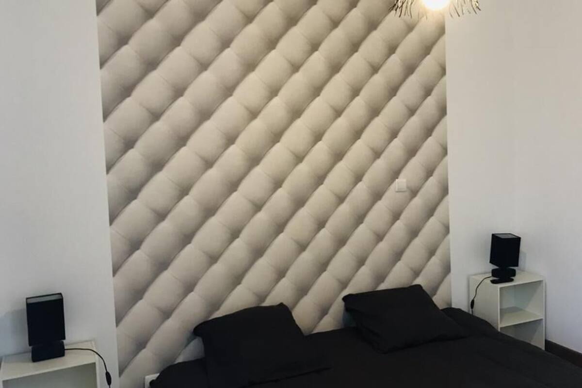 A bedroom with a tufted headboard, a bed with black bedding, a bedside table with a speaker, and a small shelf with a lamp.