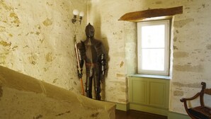 Una habitación con una escalera de madera, un ventanal con vistas y una armadura medieval apoyada en la pared.