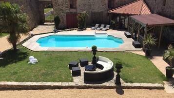 Une piscine dotée de chaises longues et d’un espace de repos couvert, entourée d’un bâtiment en pierre et de verdure.
