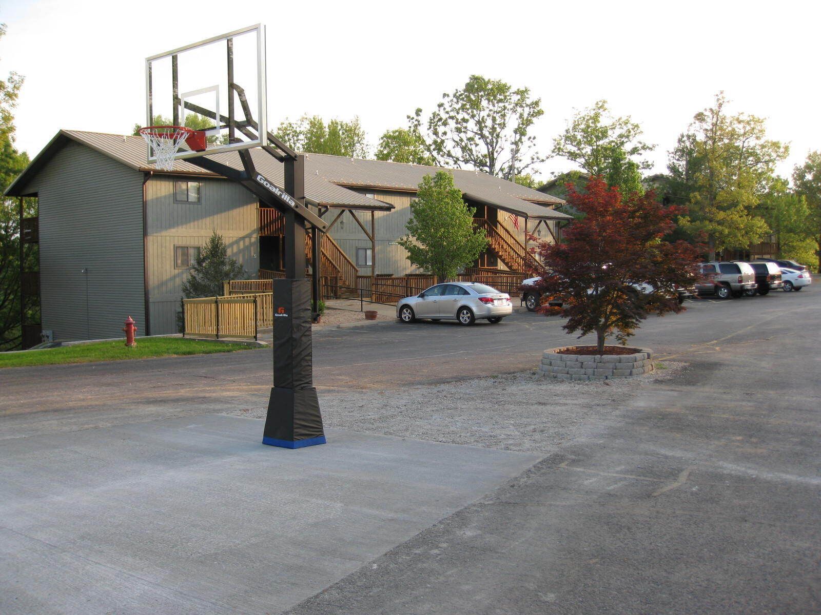 A basketball court with a hoop, a parking lot with cars, and a building with a ramp and stairs.