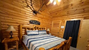 A log cabin bedroom with a wooden bed, two bedside tables with lamps, a sign above the bed, and a wooden ceiling with antlers. - 2 bed Log Cabin, w/2 Jacuzzi, 1 mi to SDC, pool, nature trails, private lake (Reeds Spring)