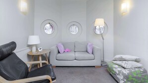 Un salon moderne avec un canapé gris, un fauteuil en cuir noir, une petite table ronde avec une lampe et un lampadaire. On y trouve également deux lampes fixées au mur et un plafond orné d’un motif de feuilles.