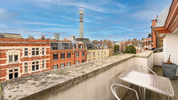 Un balcon avec une table et des chaises en métal, donnant sur un paysage urbain composé de bâtiments historiques et d’une haute tour.
