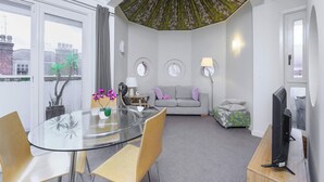 Un salon moderne avec une table à manger en verre, un canapé gris, une petite table ronde avec une lampe et un téléviseur posé sur un meuble en bois.