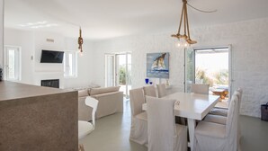 A modern living room with a white sofa, a dining table with chairs, a TV mounted on the wall, and a large window providing natural light. - White Sands Smile  · Fabulous pool villa, steps fr (Xifara)