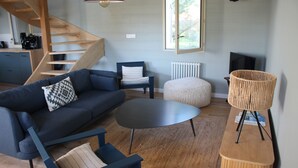 Uma sala de estar moderna com um sofá azul, uma mesa de centro redonda, uma mesinha de cabeceira de vime, um banquinho redondo pequeno, uma poltrona azul escura e uma escada de madeira.