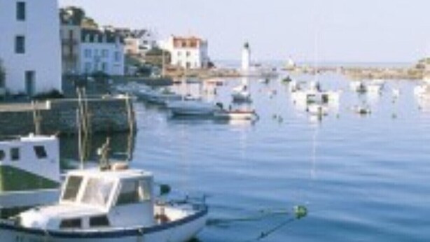 Uma marina com vários barcos atracados, um edifício branco com telhado inclinado e um farol ao fundo.
