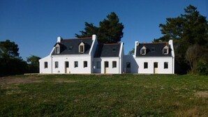 Um edifício branco com várias janelas pequenas e um telhado inclinado, rodeado por um espaço gramado e árvores.