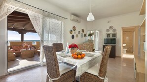 A dining area with wicker furniture, a table set with plates, a bowl of fruit, and a view of the sea through a glass door. - 1720 Casa Jonica by BarbarHouse (San Pietro in Bevagna)