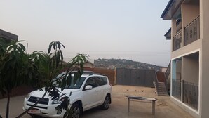 A white SUV parked in a courtyard with a building on one side and a hill in the background. - Private Executive Apartments (Accra)