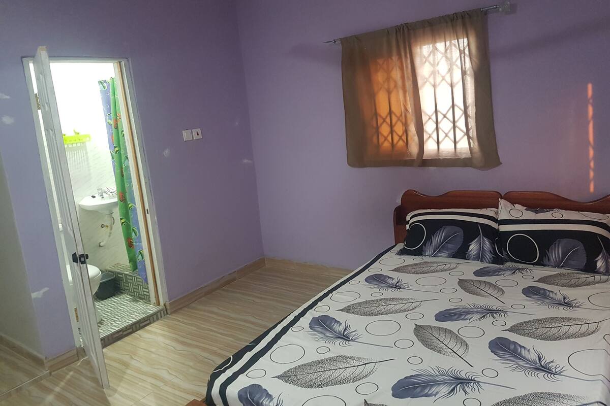 A bedroom with a bed, a window with curtains, a door leading to a bathroom, and a light switch on the wall.