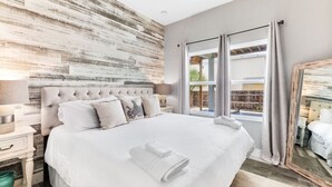 A bedroom with a large bed, a wooden accent wall, a window with curtains, and a nightstand with a lamp. - Sea Gem: Beach/Slp 10 Adults plus children/Amenities (St. Augustine)