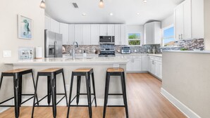 A modern kitchen with white cabinets, a large island with stools, and a view of the outdoors through a window. - Sea Gem: Beach/Slp 10 Adults plus children/Amenities (St. Augustine)