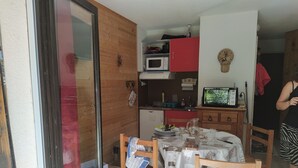Een compacte keuken met een magnetron, een rode kast en een kleine televisie. Er is een eettafel met stoelen en een krant erop.