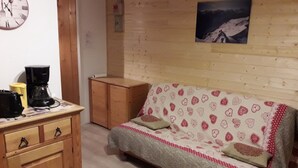 Een gezellige kamer met houten vloer, een bank met een decoratieve hoes, een houten kast, een koffiezetapparaat en een wandklok.