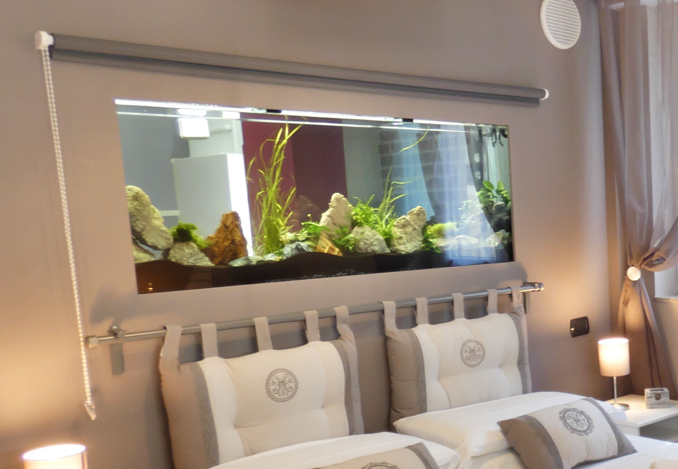 A hotel room with a large wall-mounted aquarium, a bed with two pillows, and a bedside table with a lamp.